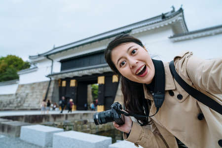 happy young lady holding camera is taking a selfie with smartphone at the entrance of Nijo Castle. asian female traveler facing to camera is excited to see the famous historic building in Japan.の写真素材