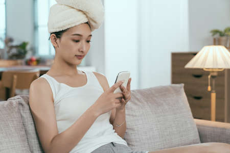 asian woman just having morning shower is using phone on sofa. pretty taiwanese lady wrapping hair...