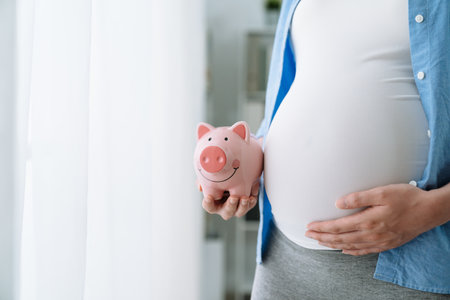 close up unrecognized asian pregnant woman holding pink piggy bank and keeping one hand on big belly. focus view on future mom abdomen with coin bank. maternity motherhood saving money concept.の写真素材
