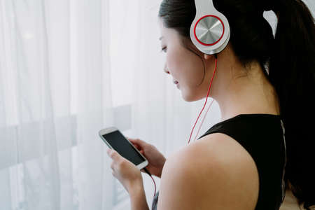 rear view korean young woman wearing headphones is turning on streaming music app. asian sportive girl watching phone screen by window is selecting songs from her playlist. technology and life.の写真素材