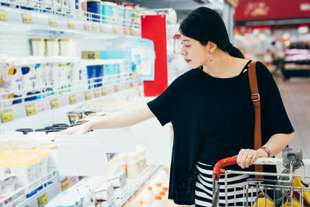 asian korean pregnant woman driving shopping cart while grocery shopping in supermarket. happy motherhood with big belly pushing trolley and stop walking. young future parent buying milk on shelf.の写真素材