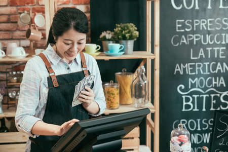 startup business successful and money concept. young woman cafe owner with dollar cash money counting earn of coffee shop on point of sale terminal machine. lady waitress laughing account on tablet.の写真素材