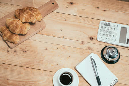 Close-up top view of cup of espresso and crispy croissant on wooden table. delicious handmade bread in coffeehouse. calculator and note for counting finance of cafe store by serving bell indoors.の写真素材