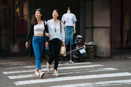 two millennial girlfriends holding hands and laughing across pedestrian crossing. full length asian charming sisters looking same side checking with cars and traffic walking on zebra road outdoors.の写真素材