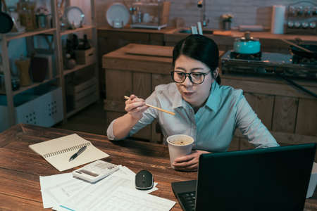 Young serious businesswoman eating chinese food from disposable paper cup and smiling enjoy sitting at wooden dining table in dark kitchen. beautiful lady worker enjoy ramen soup during overtime workの写真素材