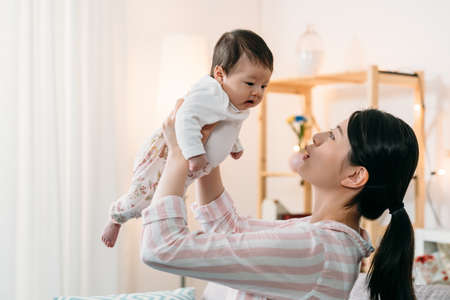 side portrait cheerful asian new mother is lifting her lovely baby daughter while playing with her in a bright living room at home.の写真素材