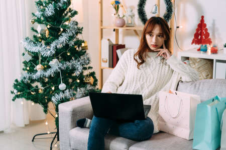 pretty asian woman looking into distance is thinking about what to buy while shopping online for Christmas gift on the computer in the living room at home.の写真素材