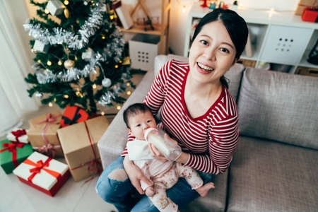 happy first-time mom looking up at camera while feeding her baby. view from above asian female seated on couch and holding her hungry child.の写真素材