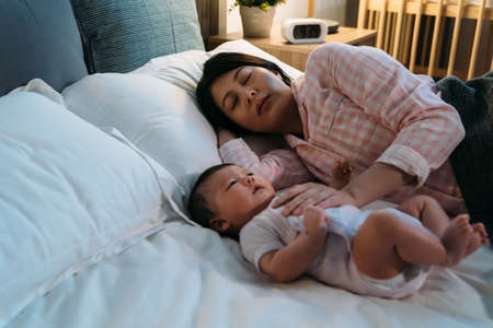 tired asian new mom wearing pajamas is falling asleep on the bed while comforting her waking baby at...