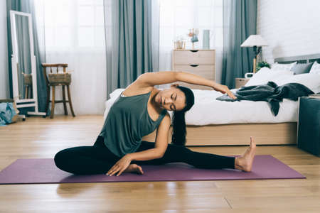 taiwanese sportive lady seated on yoga mat is warming up by doing oblique stretching in bedchamber. exercise and healthy lifestyle conceptの写真素材