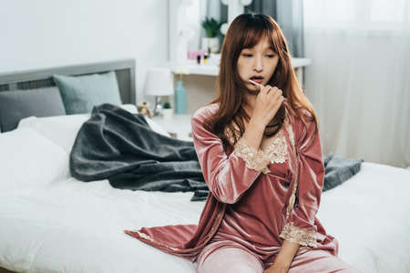 portrait asian female wearing pajamas and holding a toothbrush is cleaning her teeth with a dreamy face in the bedroom at home with morning daylight.の写真素材