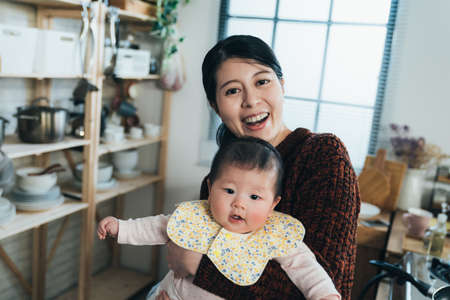 dutch angle shot joyful asian first time mom holding a lovely baby is smiling at the camera at...