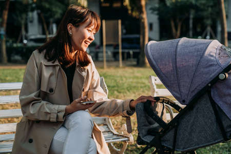 happy asian businesswoman holding a phone is playing with her baby in the stroller while resting on...