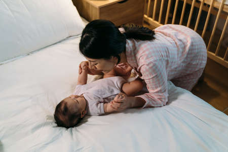 happy asian mother is leaning over and playing with her cute baby daughter near the bed before bedtime at nighttime in the bedroom at home.の写真素材