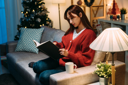 beautiful asian lady sitting with a cup of hot mulled wine is reading a book alone on the couch in a...