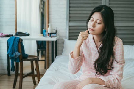 sleepy asian female sitting at bedside brushing teeth is dozing off with eyes close in the early morning at a cozy bright home bedroom interior.の写真素材