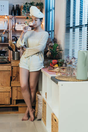 vertical full length attractive asian lady standing cross legged near window with  towel on head is sipping wine on a tranquil night after shower at home.の写真素材