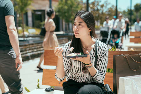 portrait beautiful asian woman is looking at the pedestrians while eating takeaway meal on the bench in downtown area on a sunny day.の写真素材