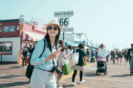 end of route 66 on santa monica pier california. happy smiling young asian chinese woman backpacker holding smartphone and camera joyful looking aside under sunshine. pretty tourist enjoy sun flareの写真素材