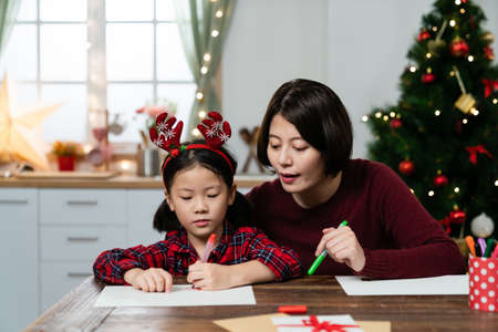 asian mother helping her young daughter making wish list of presents for Christmas at table in a...