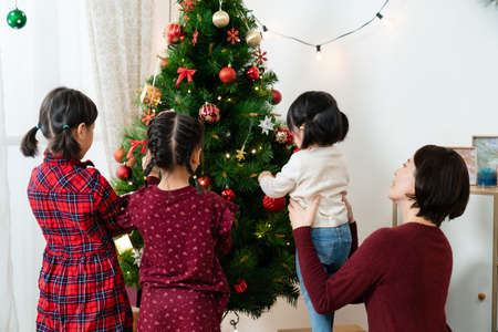 back view of an asian mother supporting her youngest daughter who is putting decoration on Christmas tree with her sibling sisters in the living room at homeの写真素材