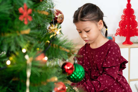 selective focus of an asian little girl preparing for xmas by a Christmas tree, putting decorations with concentration in the living room at homeの写真素材
