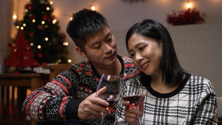 smiling asian couple in love hugging and drinking wine together while celebrating valentineâs day in a cozy home interior with Christmas decor at nightの写真素材