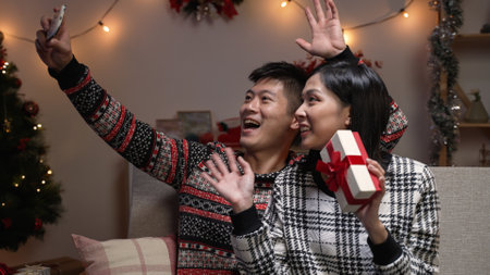 cheerful asian couple holding a Christmas gift while they are waving hi and making video call to their friends on mobile phone during Christmas holiday at homeの写真素材