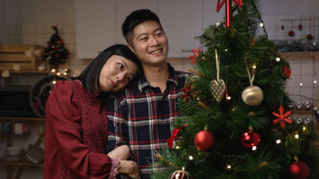 asian woman putting hanging ornament and holding her boyfriendâs arm lovingly while they are looking at decorated Christmas tree feeling happiness at homeの写真素材