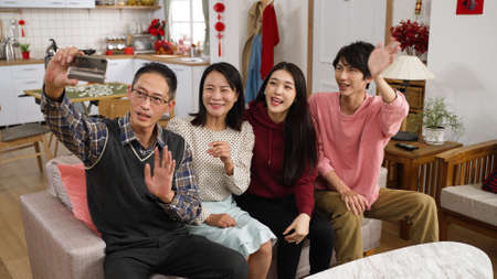 cheerful asian family waving hi to mobile phone screen while chatting with friend via video call during spring festival in living room. mother using hand gesture while talking. translation: happinessの写真素材