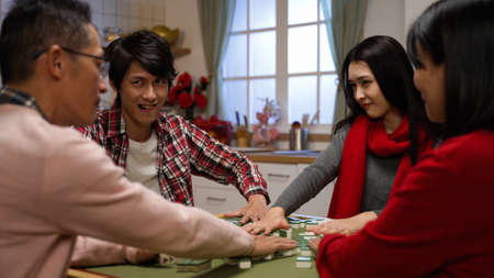 scheming son and daughter smiling at other components while mixing mahjong tiles at table. indoor family recreation for chinese lunar new year holiday conceptの写真素材