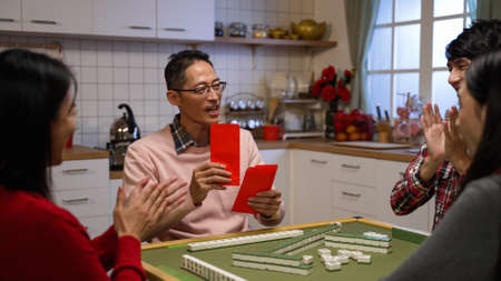cheerful father taking out from drawer and giving other family members surprise bonus red envelopes lucky money at mahjong table on chinese lunar new year's eve at homeの写真素材