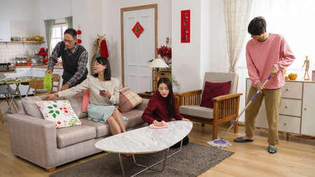 lazy asian mother playing with phone on sofa and giving command with hand gestures while other family memebers cleaning house for chinese new year. translation: spring and congratulations everyoneの写真素材