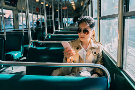 elegant businesswoman in sunglasses sending text message on mobile phone in bus. beautiful female commuter holding cup of coffee on way to work in morning. office lady using cellphone online.の写真素材