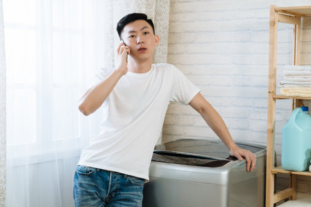 young elegant husband talking on mobile phone doing laundry at home. asian handsome guy leaning on washing machine waiting for clothes done and chatting on cellphone in bright room in apartmentの写真素材