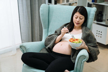 Happy asian pregnant young woman cover blanket sitting eating fruit salad on single sofa at home. elegant maternity lady with big belly enjoy vegan meal relax on couch in house living room indoorの写真素材