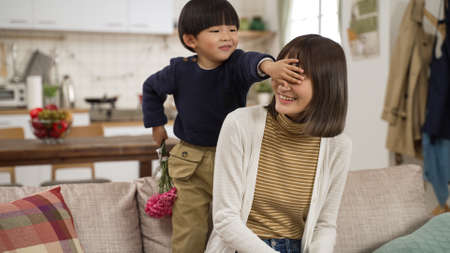 cute Asian baby kid boy covering his momâs eyes and giving her a carnation bouquet in living room on motherâs day.の写真素材