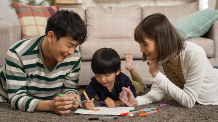 cheerful chinese family of three having fun lying prone on living room floor drawing picture together. the mother laughs at her baby sonâs silly talksの写真素材