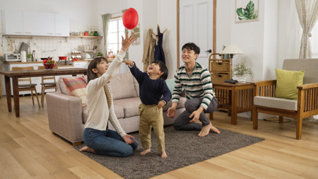 cheerful asian dad, mom and son having fun playing hit the ball game in the living room at home. they try to keep the balloon in airの写真素材
