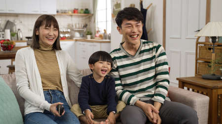 cheerful Asian family of three enjoying funny tv show together on living room couch at home. the father explaining to his son with finger pointingの写真素材