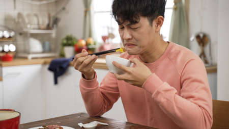 portrait of asian father making expression of disgust while tasting food with chopsticks and bowl at home dining table alone.の写真素材