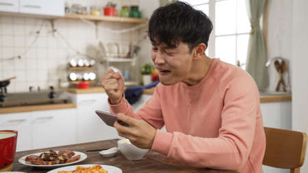 asian Japanese man playing with smartphone win the online game while eating lunch in the dining room at home. he puts down chopsticks and uses phone feeling excitedの写真素材
