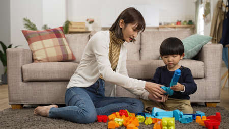 happy asian mother and baby son sitting on ground playing toy blocks together in the living room at home. she helps give him parts to buildの写真素材