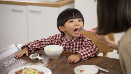 selective focus of cute Asian little boy opening his mouth as his mother feeds him a piece of meat with a fork at dining table.の写真素材
