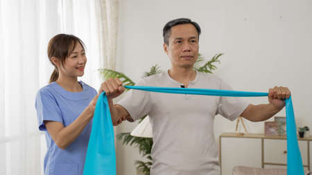 asian woman personal care attendant assisting older male working out with elastic band at home....
