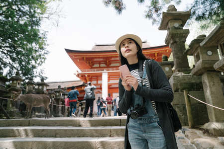 chinese girl near buddhist temple holding smartphone is looking afar. female backpacker standing on stone step is contrasting the distant view with information on phone.の写真素材
