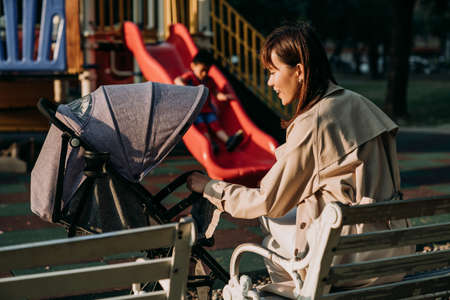 rear view joyful asian career mother is interacting with her baby in stroller while taking a break on the bench by a playground in the sun.の写真素材
