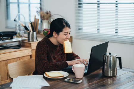 overjoyed asian housewife holding a toast with her mouth is replying letter on the computer feeling excited about income tax refund at breakfast in dining room.の写真素材