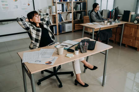 happy asian business woman is looking into distance crossing legs with smile and keeping hands behind head while relaxing on chair in open office with coworker.の写真素材
