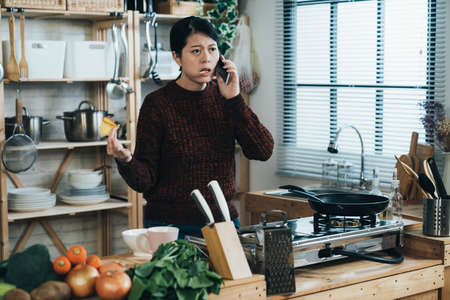 unhappy asian woman eating bread is complaining with hand gestures while talking on the phone by the kitchen stove at home in the morning.の写真素材